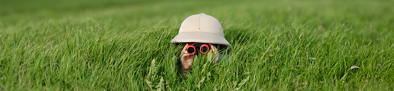 Child playing in grass field with toy binoculars and safari hat