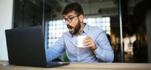 Man at laptop holding coffee with shocked expression